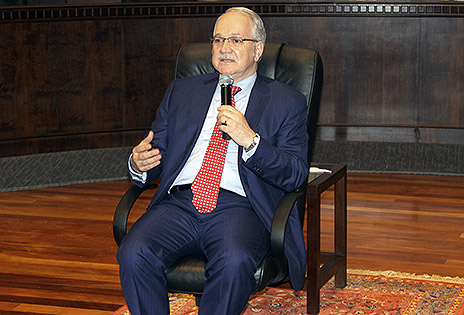 Imagem em plano americano mostra o professor Luiz Edson Fachin sentado em uma cadeira, com microfone na mão, respondendo a questão formulada por um dos magistrados presentes. O local é o plenário Pedro Ribeiro Tavares, na sede do TRT-PR. Enquanto fala, o convidado gesticula com uma das mãos.