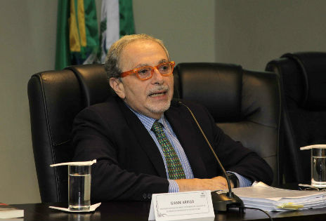 Imagem em plano médio mostra o jurista italiano Gianni Arrigo dirigindo-se, por microfone, à plateia em aula magna proferida no auditório da Escola Judicial do TRT-PR. O professor está sentado em uma poltrona, com as mãos apoiadas sobre a mesa. Uma placa de identificação colocada na mesa, à sua frente, traz o seu nome. É possível ver ainda, sobre a mesa, uma pilha de papéis em frente ao professor. Ao fundo, desfocadas, aparecem parcialmente as bandeiras do Brasil e do Estado do Paraná.