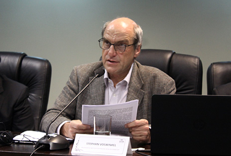 Imagem em plano médio mostra o jurista alemâo Stephan Woswinkel sentado à bancada do auditório da Escola Judicial, falando ao microfone para plateia de magistrados e servidores. O professor, usando óculos, está com um documento em mãos, como se estivesse fazendo uma leitura de algum trecho.