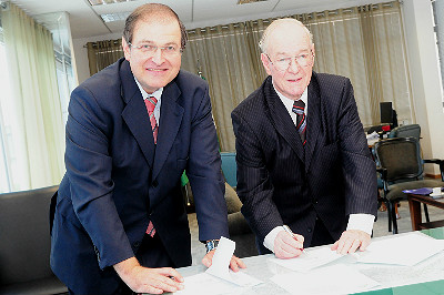 Imagem mostra o diretor da Escola Judicial do TRT-PR, desembargador Célio Horst Waldraff, e o diretor-presidente do Inter Americano, Claricio Casagrande, assinando termo de cooperação acadêmica. Os dois aparecem ligeiramente debruçados sobre a mesa, olhando para a câmera enquanto assinam o documento