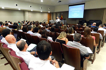 Foto em plano geral do auditório onde o evento foi realizado. A imagem traz os participantes do curso sentados enquanto o professor, de pé, na frente do auditório, fala ao microfone.