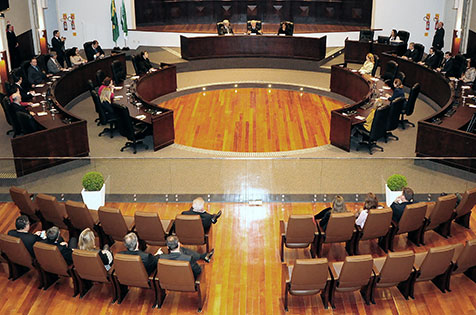 Imagem em plano geral mostra, a partir da galeria superior do plenário Pedro Ribeiro Tavares, na sede do TRT-PR, uma visão geral do ambiente em que ocorreu a sessão de leitura da ata de correição ordinária realizada no Tribunal do Trabalho do Paraná. A imagem mostra o espaço destinado aos magistrados e uma parte da plateia, com duas fileiras de poltronas centrais visíveis