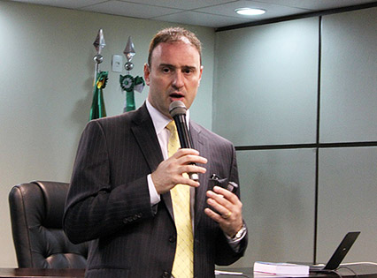 Em plano médio, o advogado André Luiz Tesser palestrando. Ele segura um microfone. Atrás do palestrante, é possível ver a mesa principal do evento, sobre a qual estão dispostos alguns objetos, como um notebook. Ao fundo, pode-se ver a ponta de dois mastros e parte das bandeiras do Brasil e do Paraná.