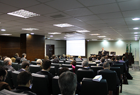 Em plano aberto, o auditório da Escola Judicial durante o evento. A foto foi tirada dos fundos do auditório. É possível ver a plateia e, ao fundo, um palestrante.