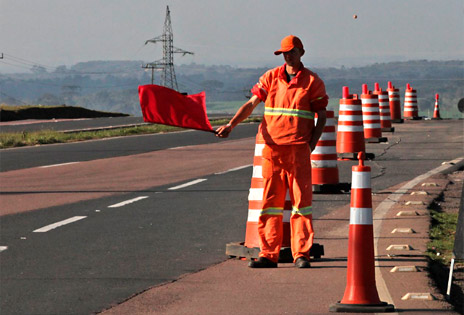 imagem mostra um operário entre cones de sinalização em uma rodovia alertando com uma bandeira vermelha para um desvio em razão de obras no trecho