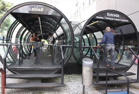 Imagem mostra estações-tubo de Curitiba e movimentação de passageiros tanto no seu interior quanto na escada de acesso para o pagamento de passagens.