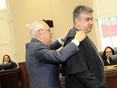 Imagem em plano médio mostra o juiz Alexandre Augusto Campana Pinheiro recebendo de seu pai a toga, durante cerimônia de posse