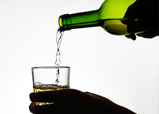 Imagem em plano fechado mostra pessoa servindo, em contra-luz, bebida alcoólica em um copo. A foto mostra apenas parte das mãos, do copo e da garrafa