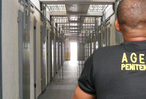 imagem ilustrativa mostra um agente penitenciário visto pelas costas, com um corredor de presídio diante dele