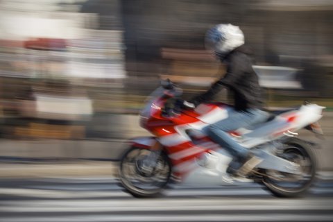 Imagem em plano aberto mostra motocilcista se deslocando em via urbana. A imagem aparece desfocada, dando sensação de movimento