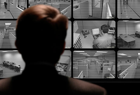 imagem ilustrativa mostra uma pessoa de costas, em primeiro plano, diante de uma tela de monitoramento de circuito interno de TV