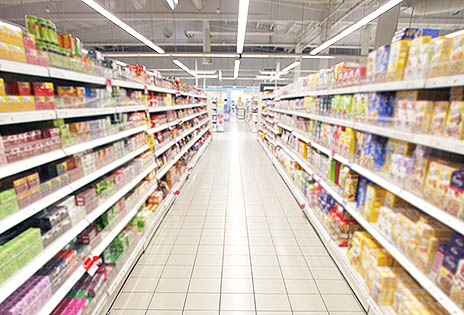 Imagem mostra corredor de supermercado, em perspectiva, com fileiras de prateleiras em ambos os lados. Não há pessoas circulando