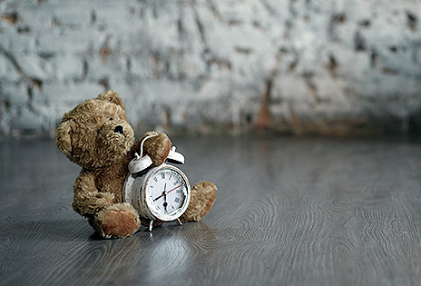 Imagem em plano médio mostra parte de um cômodo supostamente vazio, com um urso de pelúcia colocado no chão, segurando um despertador.