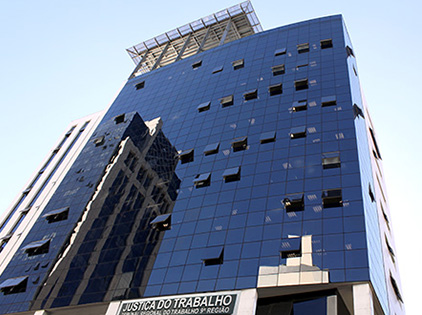 Foto em plano geral mostra a fachada do edifício-sede do Tribunal Regional do Trabalho do Paraná. A imagem foi feita em perspectiva, a partir da calçada frontal ao prédio, mostrando o perfil vertical da edificação. 