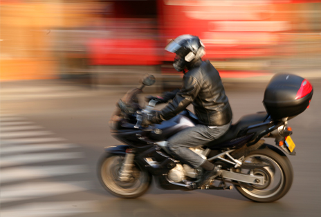 Imagem em plano geral mostra motociclista circulando em via pública. Com exceção do motociclista, toda a imagem aparece borrada, em razão de a captação ter sido feita com a câmera acompanhando o veículo em movimento.