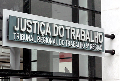 Em primeiro plano, a imagem mostra a placa da fachada do prédio sede do TRT-PR. Na placa lê-se: Justiça do Trabalho, Tribunal Regional do Trabalho da 9ª Região.