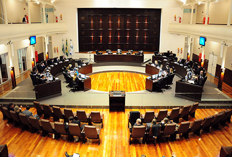 A foto, tirada de cima, em plano geral, mostra todo o Tribunal Pleno. Na parte inferior da foto, é possível ver algumas cadeiras da plateia e, no centro da foto, as bancadas do pleno, ocupadas pelos desembargadores.