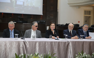 Desembargadores Ana Carolina, ao centro, Ricardo Tadeu, à esquerda na foto e Aramis, à direita, com dois dos convidados, na mesa de abertura do seminário. Eles estão sentados à mesa, coberta com longa toalha clara, tendo ao fundo parte do telão do plenário e à frente as extremidades das flores usadas na decoração, coloadas diante da mesa de honra.