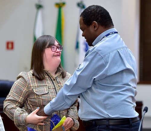 O servidor Jorge Pires, negro, de camisa azul, e a terceirizada Maria Gabriela, que tem síndrome de down, de frente um para o outro, sorriem, com as mãos dadas. 
