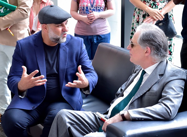 O artista Toto Lopes, homem branco com barba grisalha, est&aacute; &agrave; esquerda, fala e gesticula ao descrever a obra Nossa Terra sentado em um sof&aacute; de couro preto. Ele veste camiseta e palet&oacute; azul e utiliza uma boina cinza. &Agrave; direita, o desembargador Ricardo Tadeu Marques da Fonseca ouve o artista. O magistrado, sentado no mesmo sof&aacute;, tem a pele branca, cabelo grisalho e usa &oacute;culos escuros e veste terno cinza, camisa branca, gravata verde e usa rel&oacute;gio.