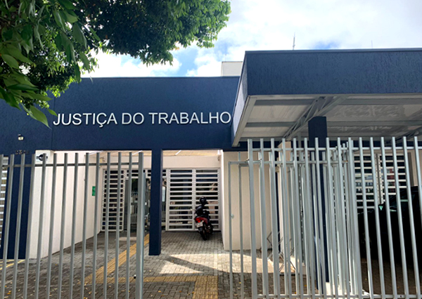 Fotografia mostra a fachada do Fórum em Francisco Beltrão. Em primeiro plano, estão grades metálicas brancas com portão de acesso. Em seguida, calçada de em paver com sinalização para pessoas com deficiência visual e, na sequência, o prédio com paredes brancas e azuis. Próximo a porta de acesso, um letreiro diz: Justiça do Trabalho. 