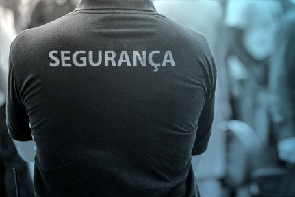 Fotografia mostra um homem de costa com camisa preta escrito segurança em um fundo desfocado. 