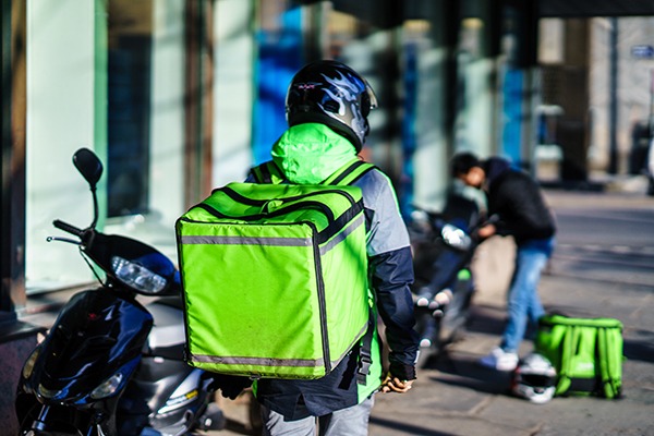 Em primeiro plano, um entregador de aplicativos de costas, com a mochila térmica na cor verde-limão à mostra. Ao lado uma motocicleta de baixa cilindrada. Ao fundo, desfocado, vê-se outro entregador de aplicativos aparentemente estacionando sua motocicleta, com a mochila no chão.