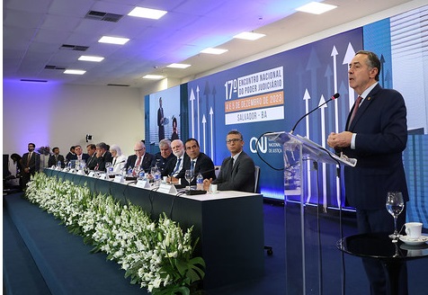 Fotografia mostra o presidente do STF, ministro Luís Roberto Barroso, discursando em primeiro plano em frente a um púlpito de acrílico com microfone. Ele veste terno escuro com gravata vermelha. Ao lado dele, uma mesa de autoridade e a fundo bannner do evento com a frase 17º Encontro Nacional do Poder Judiciário, em Salvador-BA