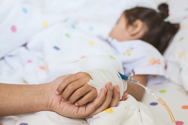 Foto mostra duas mãos em primeiro plano: a mão esquerda de um adulto que segura a mão esquerda de uma criança, que está deitada em uma cama de hospital e com acesso intravenoso com faixas sobre a mão. 