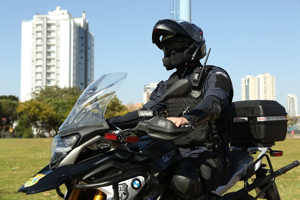 Fotografia mostra um Guarda Municipal de Ponta Grossa conduzindo uma moto em uma área verde da cidade. A fotografia tem o enquadramento acima logo acima dos pneus e mostra a moto em um ângulo lateral. Ao fundo da imagem, há um prédio. O Guarda Municipal veste uniforme da cor azul, azul capacete e balaclava.