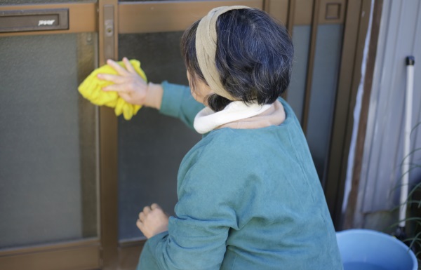 Fotografia mostra uma mulher de cócoras, de costas para a câmera, limpando uma porta de vidro com estrutura metálica. A mulher tem cabelos escuros curtos, levemente grisalhos, e usa uma faixa creme no cabelo. Ela veste uma blusa verde claro.