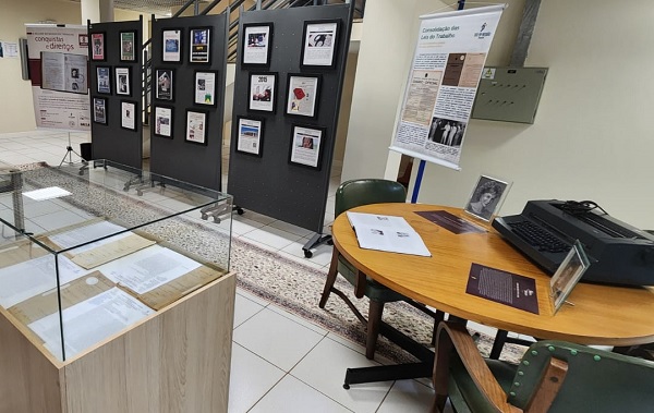 Fotografia do espaço da Mostra em Londrina. Do lado direito, uma mesa com uma máquina de escrever e fotografias antiga. Ao centro, banners e totens com a história de luta das mulheres. Do lado esquerdo, uma bancada com papeis históricos protegidos com um vidro.