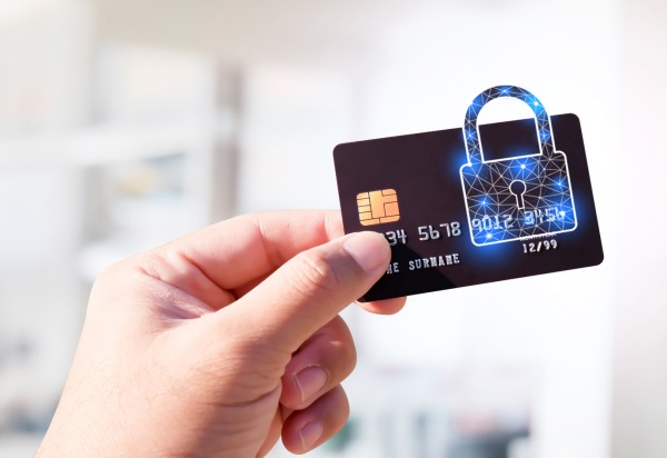 Fotografia de uma mão esquerda ao segurar um cartão de crédito azul escuro com o polegar e o indicado. Sobre o cartão de crédito, há a projeção de uma imagem holográfica de um cadeado fechado.