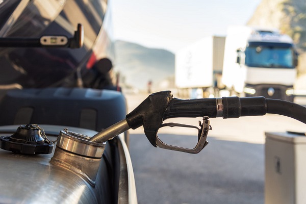 Fotografia mostra detalhe de bico de uma bomba de combustível inserido no tanque de um caminhão, que reabastece diesel em um posto. Ao fundo, desfocado, outro caminhão. 