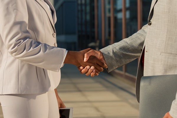 Fotografia mostra um aperto de mão entre uma mulher nega e um homem branco. Ela veste calça e blazer beges. Ele veste terno cinza claro. A imagem mostra os dois lateralmente entre o ombro e os joelhos.