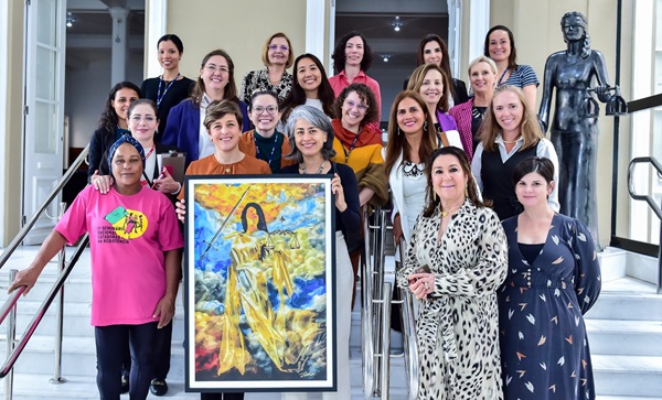 Grupo de mulheres posa para fotografia em uma escadaria. Em frente a elas, um dos quadros do artista com uma deusa com roupas predominantemente amarelas paira sob céu azul.