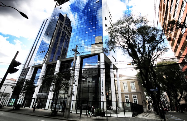 Fotografia mostra a fachada do prédio sede do TRT-PR, um edifício com pé direito alto no térreio e vidros espelhados. Imagem mostra a calçada em frente, na Alameda Dr. Carlos de Carvalho e na lateral, na rua Visconde do Rio Branco.