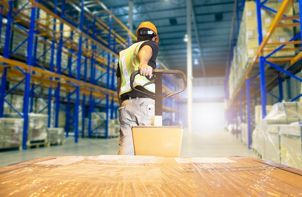 Trabalhador, de capacete e colete, puxa carrinho com caixas em armazém descarregando mercadorias de embarque de paletes,. Imagem do interior do armazém, prateleiras altas armazenamento de mercadorias em armazenagem.