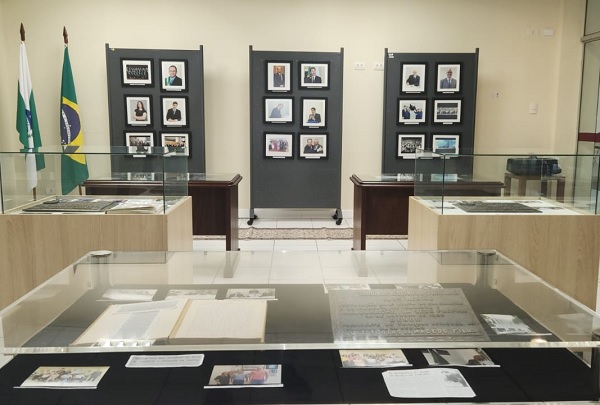 Fotografia mostra, em primeiro plano, documentos sobre uma mesa com um vidro de proteção sobre eles. Mais ao fundo, outras duas mesas de exposição no mesmo formato e no fundo da imagem três totens com fotos. 