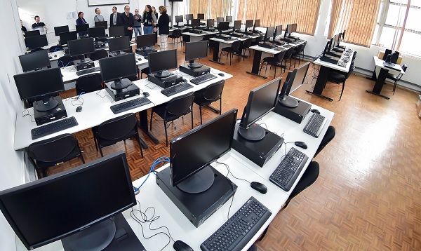 Fotografia da perspectiva do fundo de uma sala de aula com dez bancadas de mesas, cada uma com cinco computadores e perif&eacute;ricos, com cinco bancadas de cada lado, separadas por um corredeor ao centro. Ao fundo, um grupo de pessoas visita a sala. 