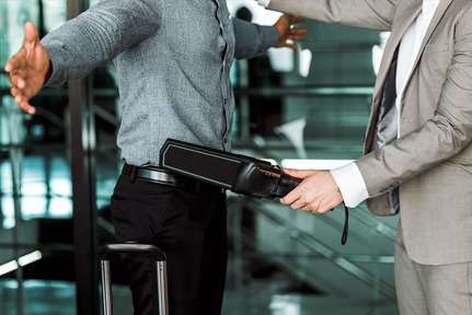 Homem de terno bege usando detector de scanner de metal, à direita, verifica outro homem, de calça preta e camisa cinza, que está de braços abertos, à esquerda. Ambos estão em pé e são mostrados da altura do joelho até o pescoço.