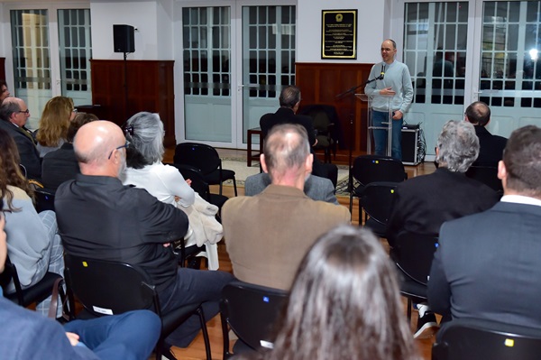 Fotografia do palestrante Caetano Galindo, homem negro, fala em frente a um púlpito. Pessoas de frente para ele e de costas para a câmera compõem a plateia.