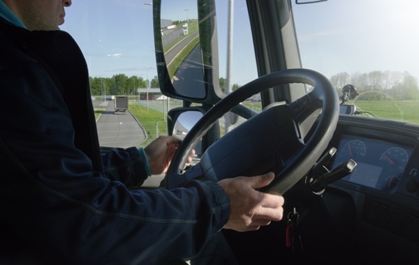 Fotografia de dentro da cabine de um caminhão. Nela, aparece do lado esquerdo parcialmente o corpo do motorista sentado com as mãos no volante de direção. À direita, o painel do veículo com a luz solar incidindo no parabrisa. Pela janela do motorista, é possível avistar uma área verde com árvores e, pelo espelho, nota-se que ele desloca-se em uma rodovia e há um outro caminhão atrás. 