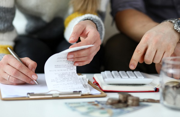 Fotografia de um casal fazendo contas juntos. A imagem está enquadrada nas mãos deles sobre uma mesa de centro. Mãos femininas escrevem em um papel do lado esquerdo enquanto uma mão masculina digita em uma calculadora do lado direito.