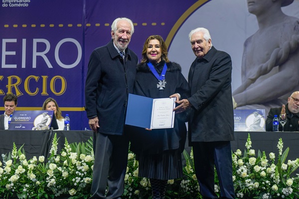 Fotografia mostra três pessoas, com a desembargadora Ana Carolina Zaina no meio entre dois homens. Ela é uma mulher branca de cabelos até a altura do ombro. Usa um colar com a homenagem de fita azul e segura um certificado. Ao lado dela, Darci Piana, homem de cabelos e barbas brancas. No outro lado, homem de cabelos brancos.