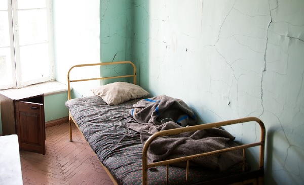 Fotografia do interior de um quarto com a pintura desgastada e uma ampla janela à esquerda. Do lado direito, uma cama de metal com colchão, coberta e travesseiro velhos e usados. Piso no local em madeira.
