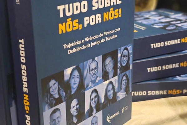 Livro Tudo sobre n&oacute;s, por n&oacute;s. H&aacute; um livro em p&eacute; e outros empilhados, sobre uma mesa. Capa azul escuro com fotos das pessoas que prestaram depoimentos &agrave; obra. 