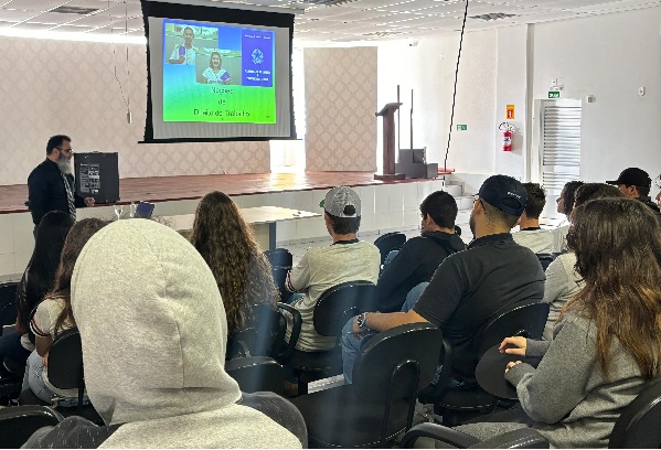 Fotografia mostra um grupo de adolescentes de costas sentados em cadeiras. Ao fundo, homem de barba grisalha está em pé e fala aos adolescentes. No centro da imagem, a uma tela de projetor com imagens de carteiras de trabalho.