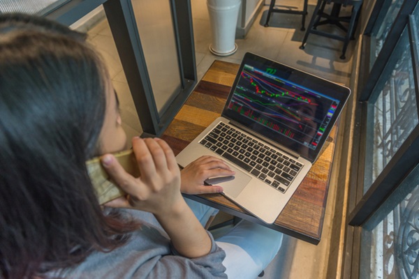 Fotografia de uma mulher de costas falando ao telefone e digitando em um computador, sentada em frente a uma mesa de madeira com computador. Na tela, imagem de gráficos.