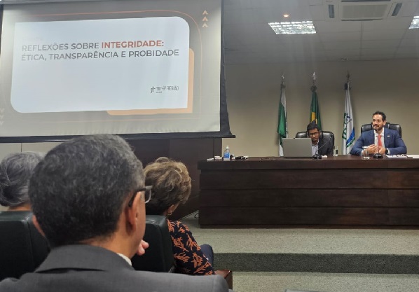 Fotografia mostra dois homens sentados em frente a bancada de madeira e, em frente, um grupo de pessoas de costas acompanha a fala. Ao lado, um projetor com uma imagem escrito: Reflexões sobre integridade: ética, transparência e probidade.